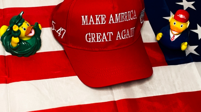 Symbolbild: Freiheitsstatue Badeente und Badeente von Donald Trump sitzen auf der amerikanischen Flagge, zwischen ihnen liegt eine Kappe mit dem Slogan "Make America great again"