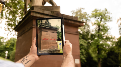 iPad filmt auf den Sockel einer Statue. Durch Augmented Reality wird eine Pinselei mit dem Text "Hitler=Krieg" von Hans Gasparitsch (1918-2002) sichtbar.