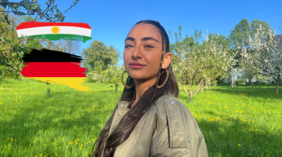 Junge Frau mit dunkelbraunen Haaren und grünen Augen, stolzer Blick in die Kamera. Nebendran sind zwei Flaggen, einmal die deutsche und kurdische.