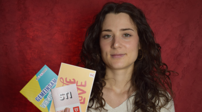 Protagonistin Lena Dillmann steht vor einem roten Hintergrund, schaut in die Kamera und hält Aufklärungsmaterial in der Hand.