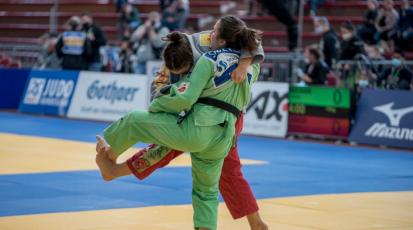Eine Frau in grünem Judoanzug und eine Frau in grauer Kutte und roter Judohose kämpfen auf blauen und gelben Judomatten. 