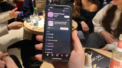 Eine Hand hält ein Handy. Darauf zu sehen ist ein Chat namens Stuttgart Friends, im Hintergrund sieht man Menschen in einer Bar.