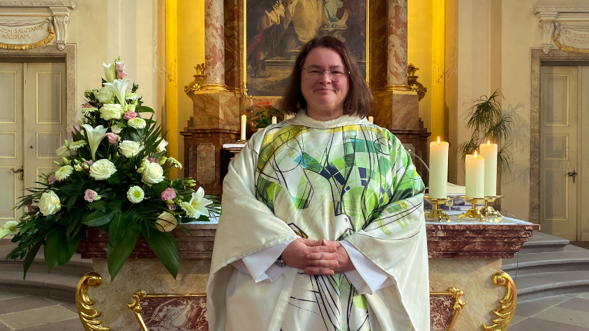 Pfarrerin Sabine Clasani steht vor dem Altar. Sie trägt ein weiß-grünes Gewand und lächelt.