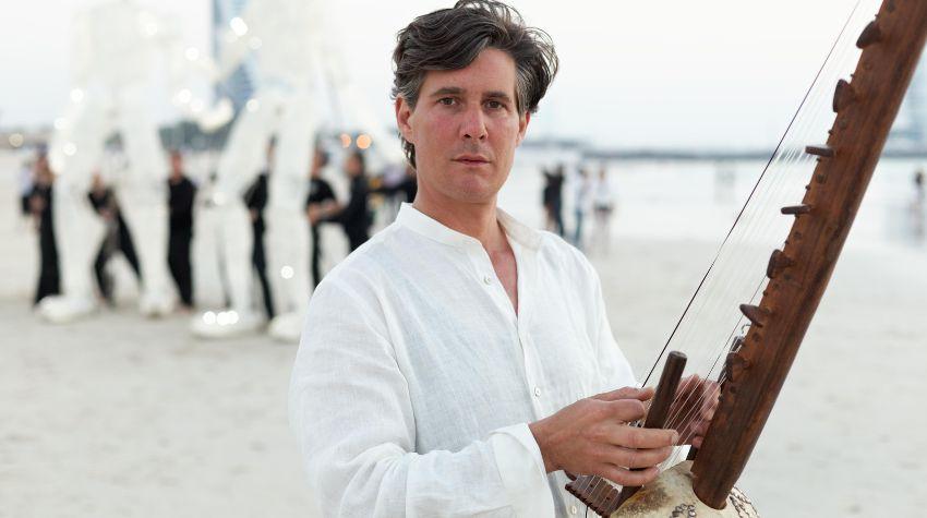 Stefan Charisius befindet sich an einem Strand und hält die westafrikanische Harfe Kora in der Hand. Im Hintergrund sind zwei Großpuppen mit jeweils fünf Puppenspielenden zu sehen.