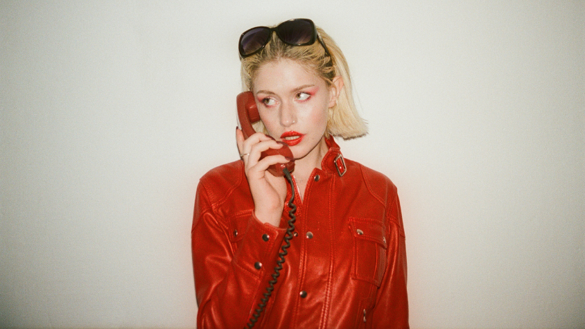 Yeva in einer auffälligen Roten Jacke hält ein rotes Kabeltelefon an ihr Ohr.