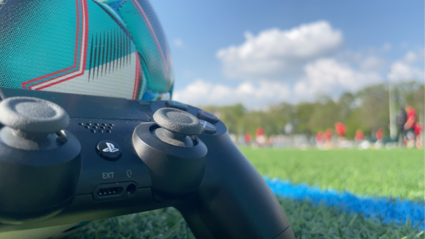 Ein Controller liegt vor einem Fussball. Im Hintergrund kann man unscharf eine Fußballmannschaft sehen.