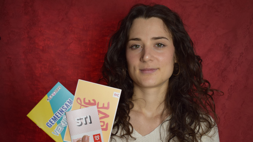 Protagonistin Lena Dillmann steht vor einem roten Hintergrund, schaut in die Kamera und hält Aufklärungsmaterial in der Hand.