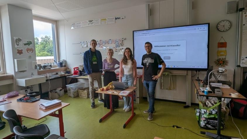 Die Studierenden und ein Grundschullehrer stehen gemeinsam vor einer digitalen Tafel, auf der die WebApp zu sehen ist.