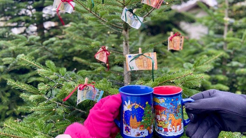 Geldscheine hängen an einem Weihnachtsbaum und zwei Weihnachstmarkttassen stoßen im Vordergrund an. 