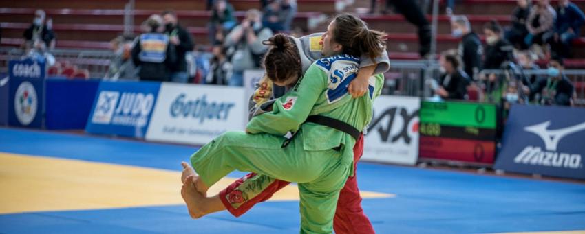 Eine Frau in grünem Judoanzug und eine Frau in grauer Kutte und roter Judohose kämpfen auf blauen und gelben Judomatten. 