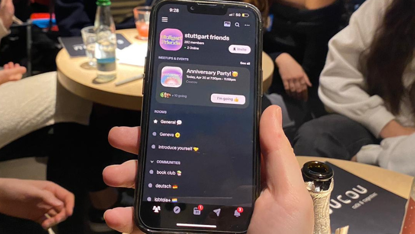 Eine Hand hält ein Handy darauf zu sehen ist ein Chat namens Stuttgart Friends, im Hintergrund sieht man Menschen in einer Bar