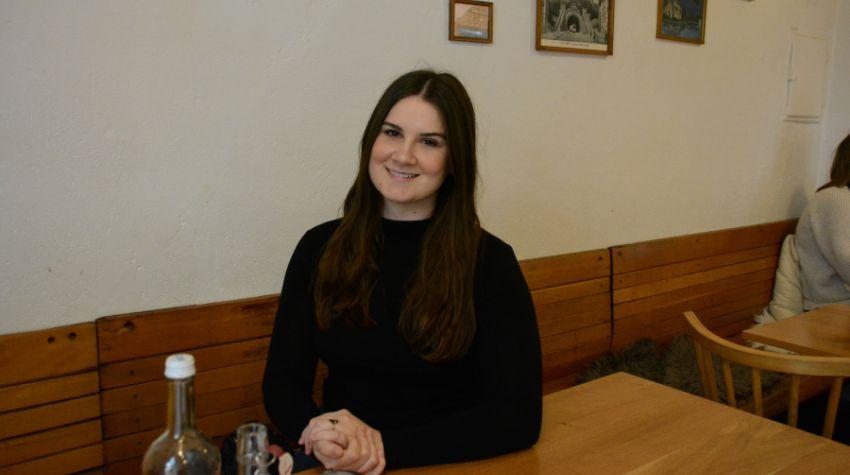 Zu sehen ist eine Frau mit dunklen langen Haaren die an einem Tisch sitzt und in die Kamera lächelt