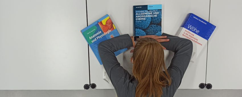 Eine Frau hat den Kopf auf ihren Händen abgelegt. Unter ihren Armen liegen ein Lehrbuch mit dem Titel "Anatomie und Physiologie", ein Lehrbuch mit dem Titel "Allgemeine und Anorganische Medizin" und ein Lehrbuch mit dem Titel "Einführung in die allgemeine Betriebwirtschaftslehre". 