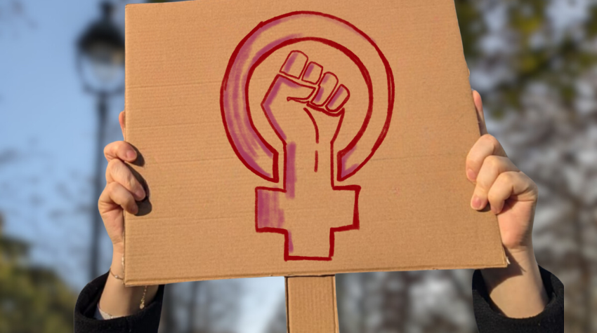 Pappschild mit feministischem Symbol: Erhobene Faust in einem Venuszeichen, gehalten von Händen vor unscharfem Hintergrund. Symbol für den Kampf um Gleichberechtigung und Sichtbarkeit von Frauen.