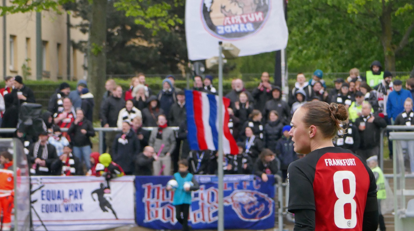 Im Vordergrund ist eine Spielerin der Eintracht Frankfurt Frauen während eines Spieles im Stadion am Brentanobad zu erkennen, im Hintergrund zu sehen sind eine Fahne und mehrere Banner des FFC-Fanclubs Nutria-Bande, welcher den Verein bei allen Heim- und Auswärtsspielen unterstützt.