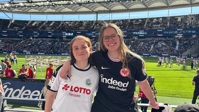 Zwei Fans stehen auf der Tribüne im Deutsche Bank Park und halten sich Arm in Arm - eine trägt das alte Trikot des 1. FFC Frankfurt und eine das neue Trikot der Eintracht Frankfurt Frauen. Im Hintergrund sind der Rasen und die gefüllten Tribünen des Deutschen Bank Parks zu sehen.