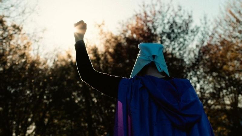 Eine Frau in lila Robe und weißer Haube richtet ihre Faust nach oben