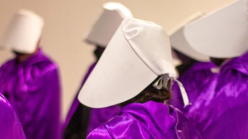 Frauen in lila Roben und weißen Umhängen stehen in einem leeren Raum