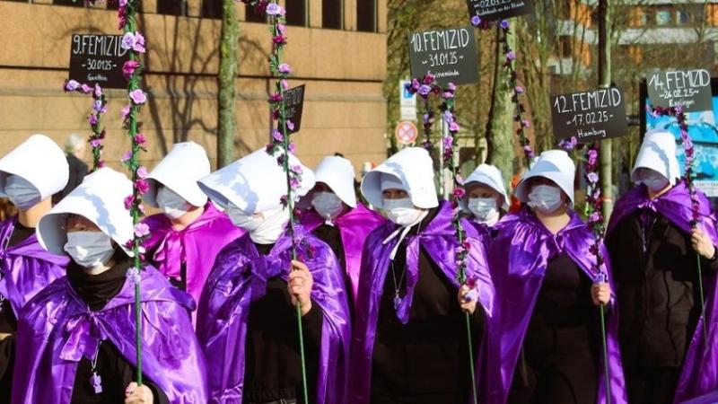 Eine Gruppe aus Frauen in lila Umhängen und weißen Hauben läuft mit Protestschildern zu Femiziden durch eine Straße