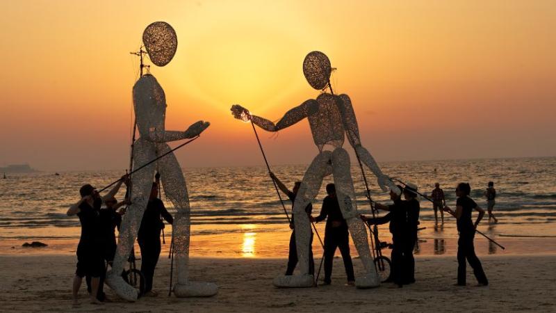 Zwei Großpuppen stehen am Strand und werden von jeweils fünf Puppenspielenden zum Leben erweckt. Hinter den Puppenspielenden und den Zuschauenden geht die Sonne im Meer unter.