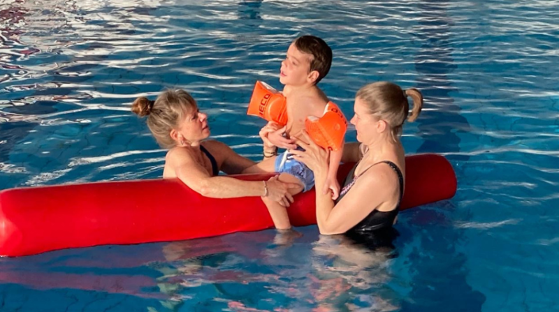 Junge sitzt auf einer roten Stange im Wasser und wird von seiner Mutter und Annette gestützt