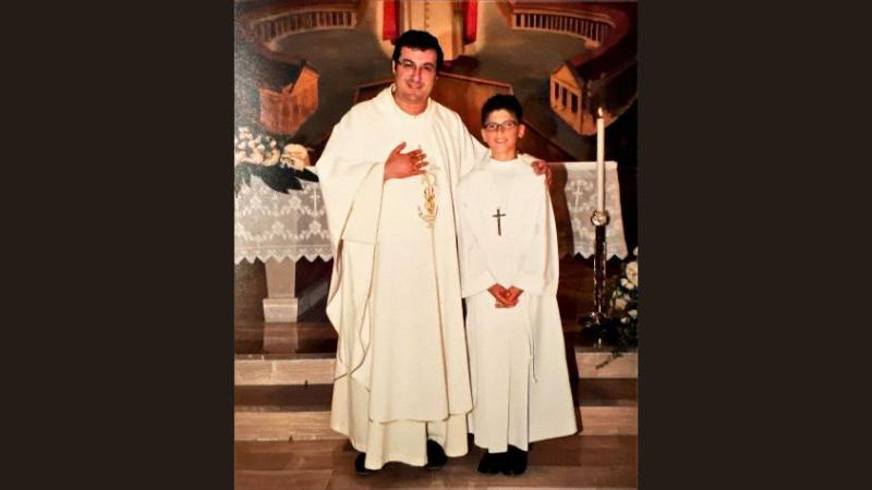 Eine Kind steht neben einem Pfarrer vor dem Altar in einer Kirche, beide tragen liturgische Gewänder.