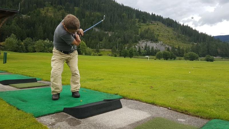 Marcel Richter steht auf einer Golf-Abschlagplattform. Er ist kleinwüchsig und trägt ein graues T-Shirt und eine beige Hose. Er hält einen Golfschläger in seinen Händen. Rechts von ihm erstreckt sich der Golfplatz. Oben rechts vor dem Himmel kann man den Ball sehen, den er gerade geschlagen hat.