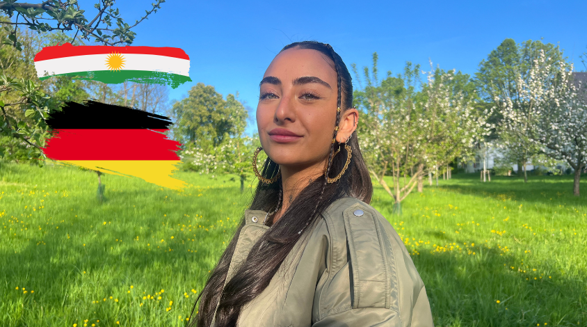 Junge Frau mit dunkelbraunen Haaren und grünen Augen, stolzer Blick in die Kamera. Nebendran sind zwei Flaggen, einmal die deutsche und kurdische.