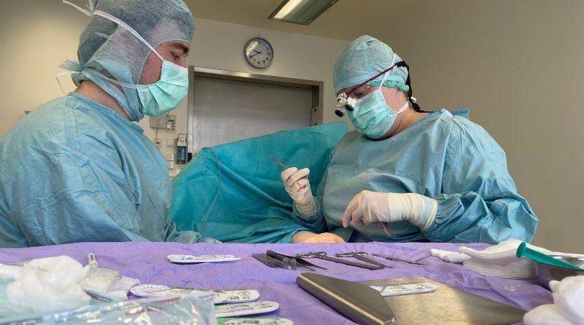 Zwei Chirurg*innen sitzen in sterilen Kitteln an einem OP-Tisch und operieren an einer Hand, im Vordergrund steht ein Tisch mit Instrumenten